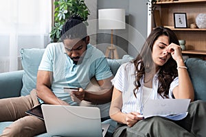 Focused worried couple paying bills online on laptop with documents sitting together on sofa at home, serious confused man and