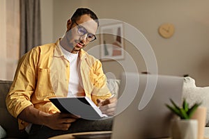 Focused young African American man using laptop, working online, taking notes during business meeting at home