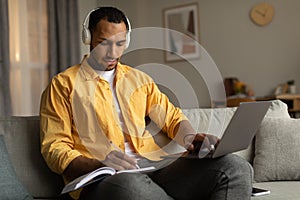 Focused young black man in headphones having online business conference on laptop, taking notes at home