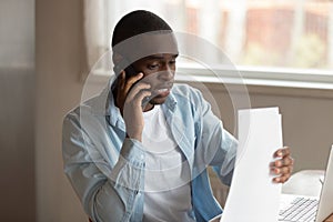 Focused young black man holding documents, calling to financial advisor.