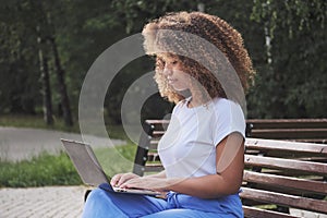 Focused young black woman student preparing for exams using laptop and sitting on park bench.