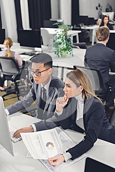 Focused young businessman and businesswoman using desktop computer and working with papers