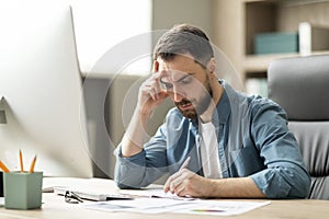 Focused young businessman sitting at desk in office and writing in notepad