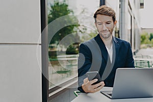 Focused young businessman working on laptop in outdoor cafe and browsing phone notifications