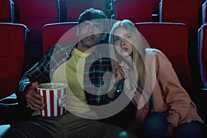 Focused young couple, attractive man and woman sitting at the cinema, watching a movie with enthusiasm and holding hands