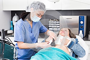 Focused young female cosmetologist in a protective mask makes an elderly woman client a vacuum massage of the abdomen