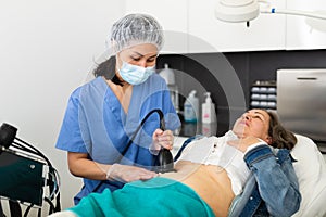 Focused young female cosmetologist in a protective mask makes an elderly woman client a vacuum massage of the abdomen