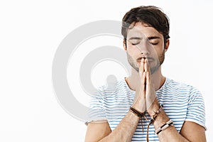 Focused young hopeful hispanic guy with beard in striped t-shirt holding hands in pray over lips standing with closed