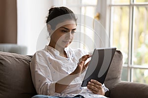 Focused young Indian tablet user woman relaxing on sofa