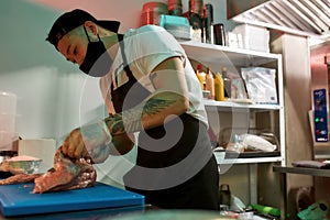 Focused young male cook wearing protective mask and gloves slicing raw pork meat on a plastic board while working in