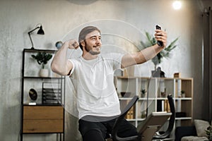 Focused young male wearing sportswear doing selfie, showing biceps while using exercise bike.