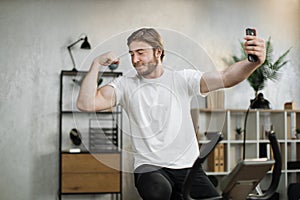 Focused young male wearing sportswear doing selfie, showing biceps while using exercise bike.