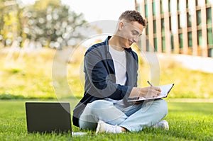 A focused young man sitting on the grass in a park, writing in a notebook with a laptop