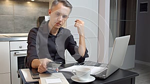 Focused young man working on laptop and using smartphone while sitting in kitchen at home