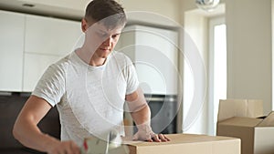 Focused young man wrapping cardboard boxes with sticky tape to move in new flat. Male packing up furniture and home