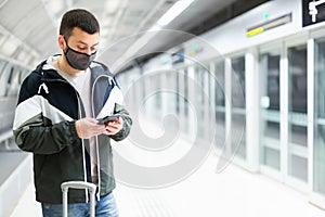 Traveler in mask standing with suitcase and phone on subway station platform