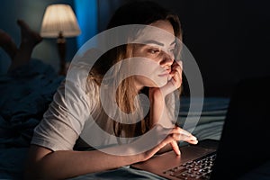Focused young woman looking at laptop screen working on project until night, lying on bed with laptop. Girl freelance IT