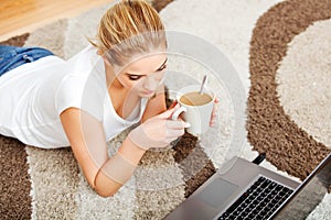 Focused young woman using laptop while lying on floor and drinking coffee