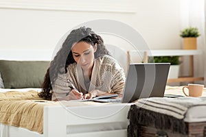 Focused young woman using laptop, working or studying online, taking notes during business meeting or webinar at home