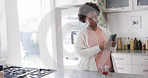 Focussed african american woman using smartphone standing in kitchen at home, slow motion