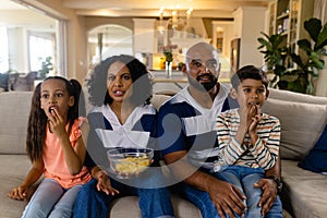 Focussed biracial family sitting on couch eating snacks and watching tv