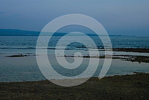 Foggy blue remote shore with pink haze, dark shore and calm sea on sunset, low tide. Tropical indonesian landscape with islands.