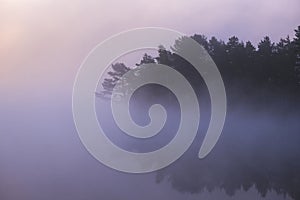 Foggy morning on the shore of a forest lake in early autumn. Trees drown in blue haze