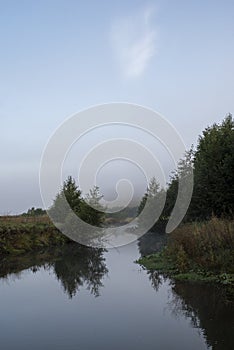 Foggy morning on a small river. Reflection of trees on the water surface