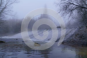 Foggy winter landscape, river with water falling down a waterfall, rocks and bare trees. Long exposure photo