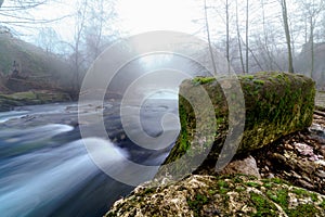 Foggy winter landscape, river with water falling down a waterfall, rocks and bare trees. Long exposure photo