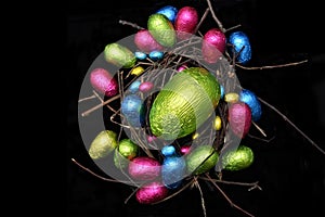 Foil wrapped multi coloured easter eggs in pink, green, blue and yello in a natural nest made of wood, sticks and twigs.