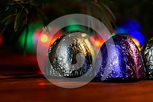 Foil-wrapped sweets lie on a wooden surface amid a Christmas tree and multi-colored LED lights. Night magic. Macro. Bokeh, volume