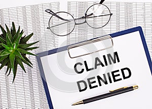 Folded business papers, pen, glasses, a flower in a pot and a tablet with a sheet of paper with the text CLAIM DENIED on the