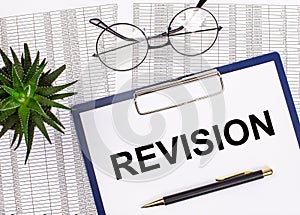 Folded business papers, pen, glasses, a flower in a pot and a tablet with a sheet of paper with the text REVISION on the desktop.