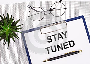 Folded business papers, pen, glasses, a flower in a pot and a tablet with a sheet of paper with the text STAY TUNED on the desktop
