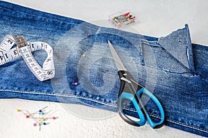 Folded in half blue jeans with large hole on pant leg below the knee, sewing pins, tailor tape and scissors