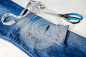 Folded in half blue jeans with a large hole on pant leg below the knee, sewing pins, tailor tape and scissors