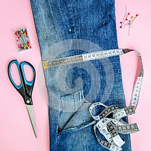 Folded in half blue jeans with large hole on pant leg, sewing pins, tailor tape and scissors