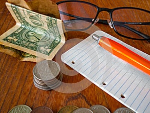 Folded One US dollar banknote, stack of coinsUS quarter dollar on the top, a notepad with a pen, glasses and a pile of coins in