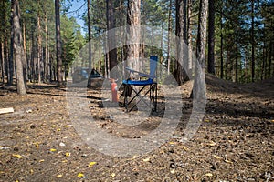 A folding camping chair is located in a clearing in a wild forest on a sunny summer day next to a fire extinguisher to