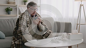 Folk medicine. Sick senior man with fever wrapped in warm plaid drinking hot tea, suffering from headache