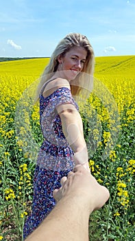 Follow me. Blonde girl in dress with flower print on the blooming yellow rapeseed field