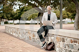 Following up on a hot lead. a mature businessman sitting on a wall and using a laptop against a city background.