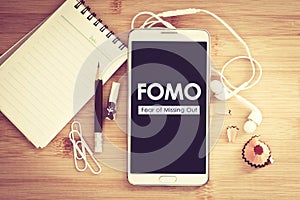 FOMO , Fear of missing out text on  Smart phone with a Blank notepad and  pencil  on wooden table
