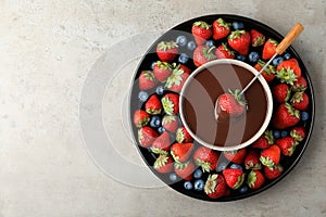 Fondue fork with strawberry in bowl of melted chocolate surrounded by different berries on light table. Space for text