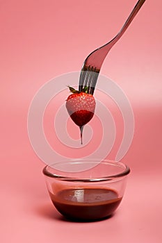 Fondue strawberry soaked in hot black chocolate on a fork with pink background with minimalist style