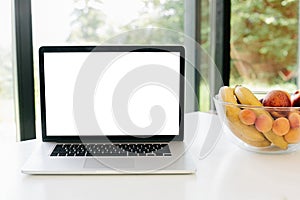 Food blog. Laptop with blank white screen and fruits in glass bowl on countertop   in modern kitchen with big windows, copy space