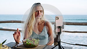 Food blogger woman films video about green salad bowl, dieting for fit skinny body at cafe, sea view. Travel influencer