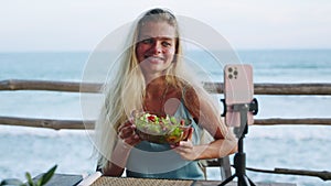 Food blogger woman films video about green salad bowl, dieting for fit skinny body at cafe, sea view. Travel influencer