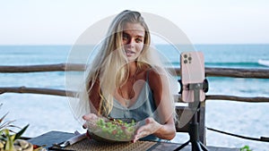 Food blogger woman films video about green salad bowl, dieting for fit skinny body at cafe, sea view. Travel influencer